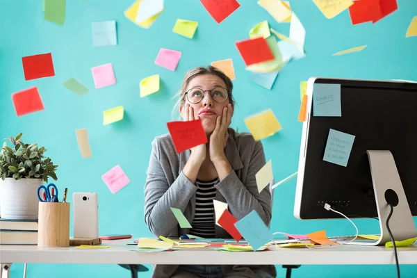 Stressad ung affärskvinna som tittar upp omgiven av post-its på kontoret. — Stockfoto