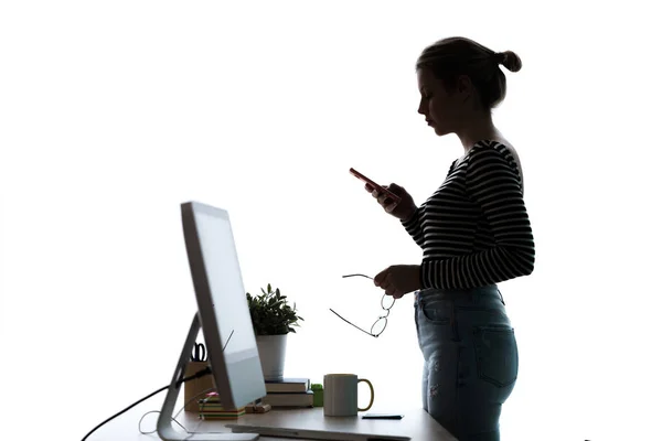 Ganska ung kvinna använder sin mobiltelefon när du står framför dator över vit bakgrund. — Stockfoto