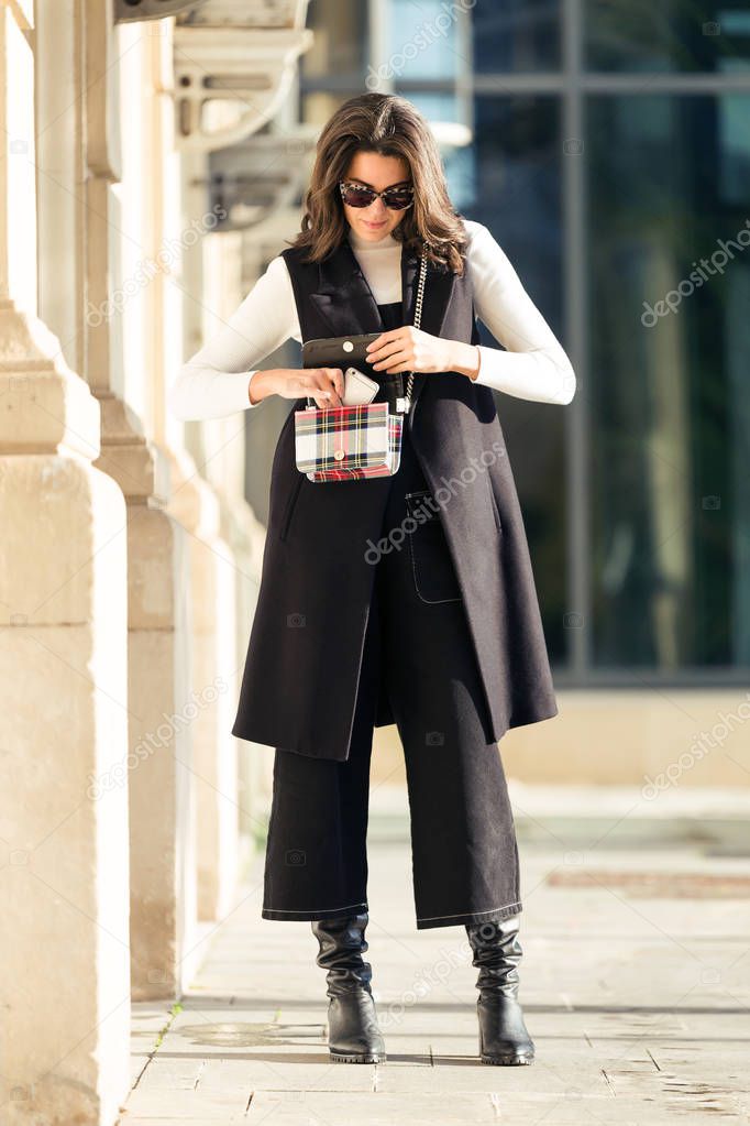 Shot of pretty young woman taking out the phone from the bag while standing in the street.