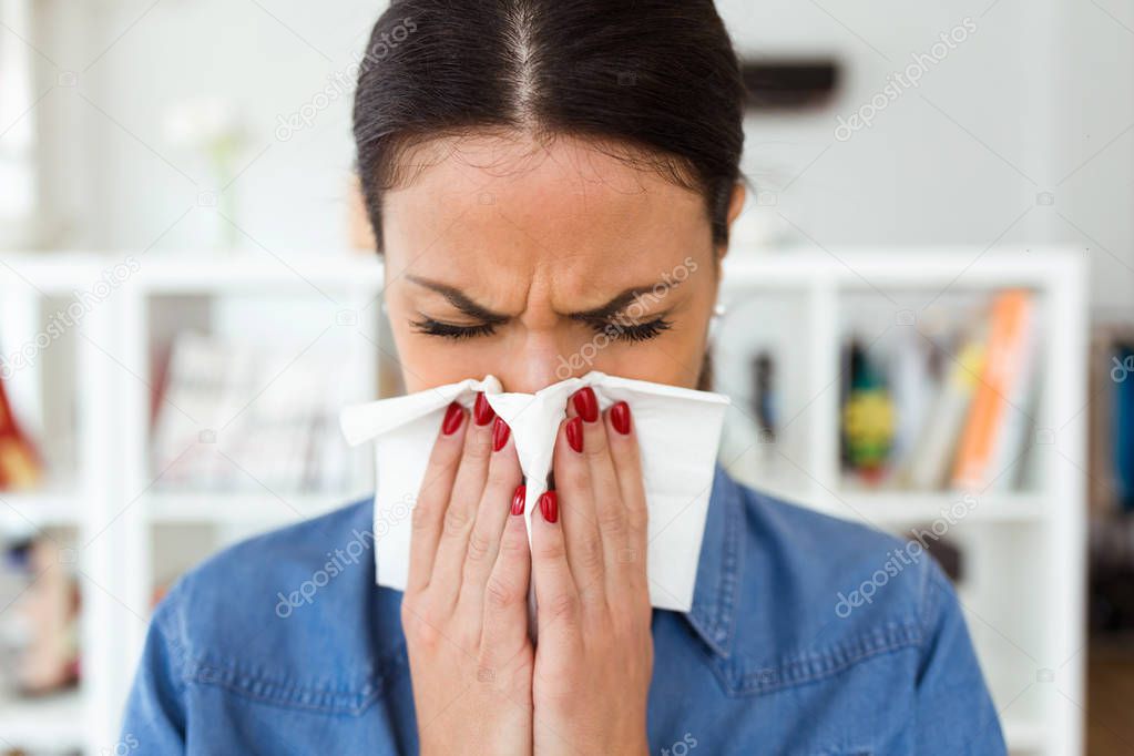 Shot of unhealthy young woman sneezing in a tissue in the living room at home.