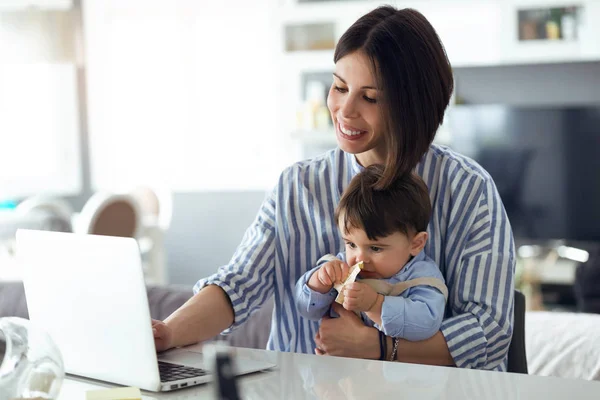 かなり若い母彼女の赤ちゃんを自宅に彼女の足の上に座ってノート パソコンで作業のショット — ストック写真