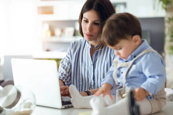 Shot of pretty young mother working with laptop while her baby sitting on the countertop in the kitchen at home.