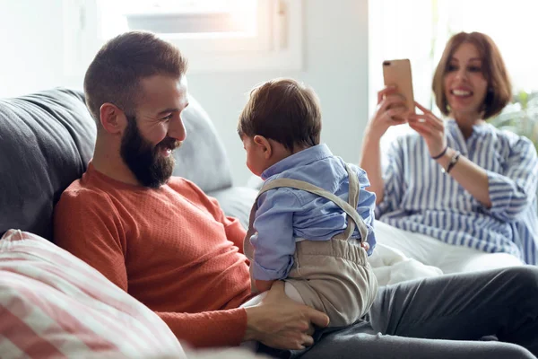 ハンサムな若い父親母親自宅のソファーには それらの写真を撮っている間彼の赤ちゃんと遊ぶことのショット — ストック写真