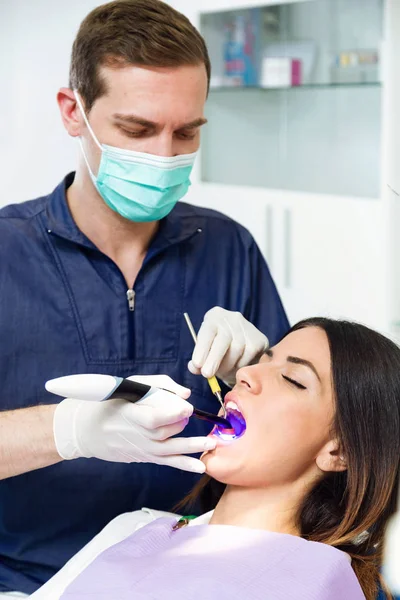 Shot of handsome confident dentist whitening tooth to female patient on dental chair in dental clinic.
