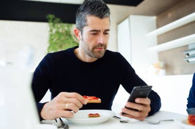 Yakışıklı genç adam aile evde mutfakta kahvaltı tadını çıkarırken onun cep telefonuyla çekim.