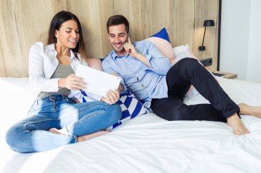 Mutlu genç çift otel odasında yatağın üstünde otururken dijital tablet ve cep telefonu kullanarak çekim.