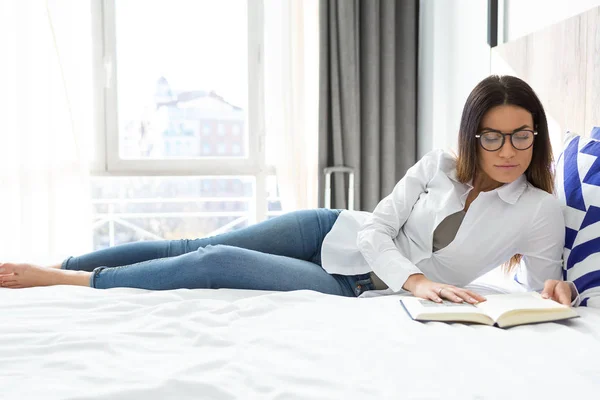 ホテルの部屋でベッドの上に横たわっている間本を読んでかなり集中して若い女性のショット — ストック写真
