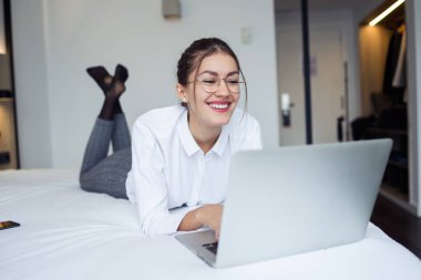 Otel odasında yatakta yatarken onu laptop ile çalışan genç iş kadını gülümseyen in atış.