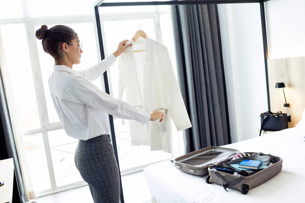 Shot of elegant young businesswoman in suit unpacking her suitcase on bed in hotel room.