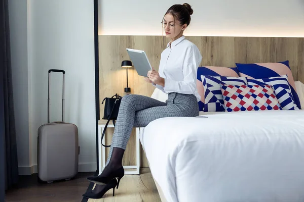 Shot of attractive young businesswoman using her digital tablet sitting on the bed at hotel room.