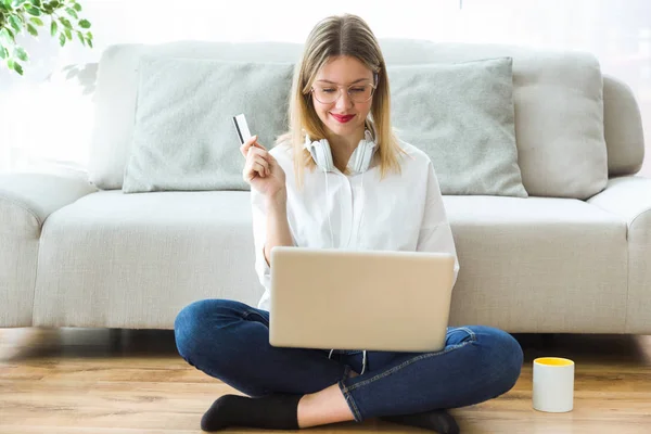 家の床に座ってコンピューターとオンライン ショッピングのための白のクレジット カードを保持している幸せな若い女のショット — ストック写真