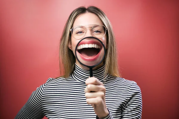 Funny young woman showing her mouth trought magnifying glass over pink background. — Stock Photo, Image