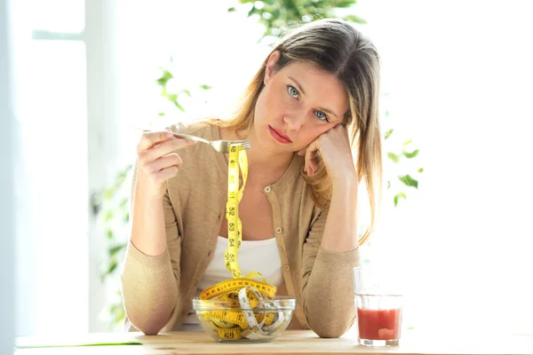 Worried beautiful woman holding fork with measuring tapes sitting in living room at home. — Stock Photo, Image