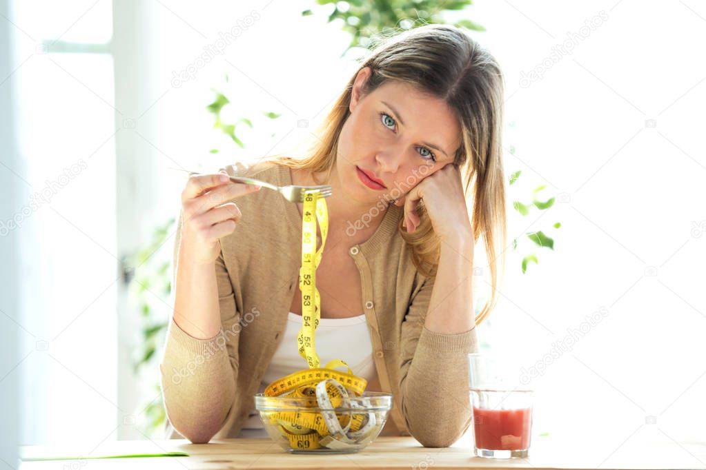 Worried beautiful woman holding fork with measuring tapes sitting in living room at home.