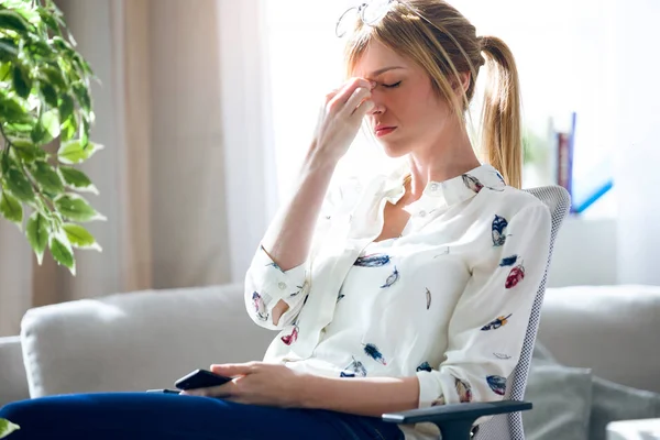 Trött ung kvinna med huvudvärk håller sin mobiltelefon på kontoret. — Stockfoto