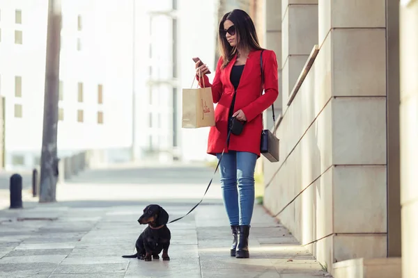 Młoda kobieta moda spaceru z psem mały podczas pisania wiadomości SMS z jej smartphone na ulicy w mieście. — Zdjęcie stockowe