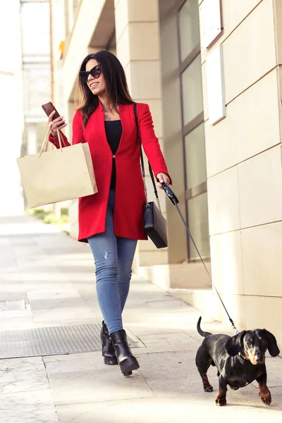 Mujer joven con estilo paseando con su perrito mientras envía mensajes de texto con su teléfono inteligente en la calle en la ciudad . — Foto de Stock