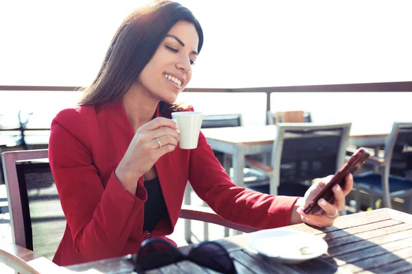 Jovem alegre bebendo café enquanto mensagens de texto com seu telefone celular na mesa wodden no terraço de um café . — Fotografia de Stock
