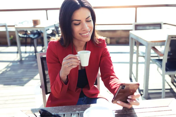 Fröhliche junge Frau trinkt Kaffee, während sie mit ihrem Handy auf dem Holztisch auf der Terrasse eines Cafés textet. — Stockfoto