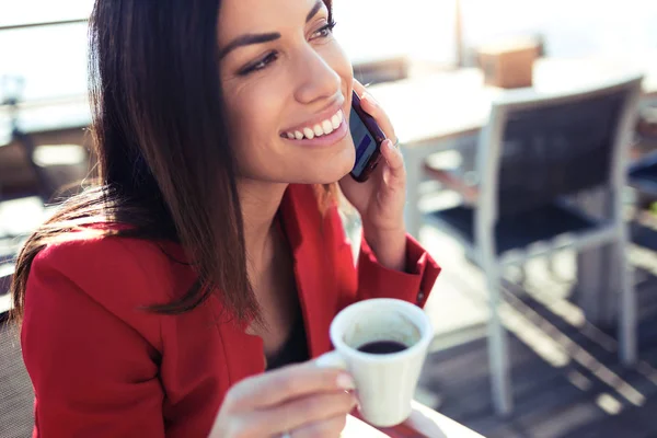 Sorridente giovane donna che beve caffè mentre scrive con il suo telefono cellulare sul tavolo di wodden nella terrazza di una caffetteria . — Foto Stock