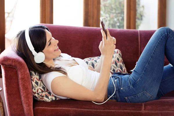 Mooie jonge vrouw luisteren naar muziek met een koptelefoon en het gebruik van mobiele telefoon, terwijl rusten op een bank op hom — Stockfoto