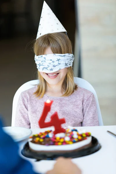 Menina bonita feliz sentado na mesa com olhos de cobertura enquanto espera bolo de aniversário na cozinha em casa . — Fotografia de Stock