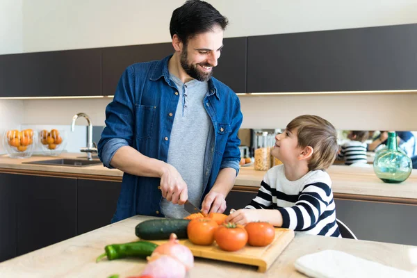 Atraente jovem pai e seu pequeno filho bonito cortando legumes em mesa de madeira na cozinha em casa . — Fotografia de Stock