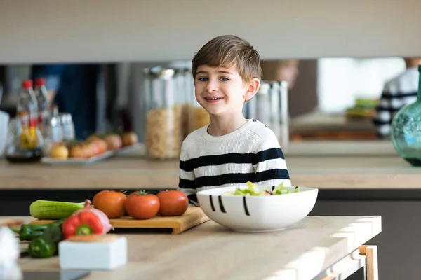 Menino bonito cortando legumes na mesa de madeira enquanto olha para a câmera na cozinha em casa . — Fotografia de Stock