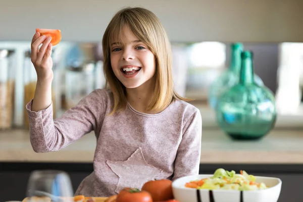 Mooie schattige kleine meid bereidt salade in een kom terwijl het tonen van tomaat aan camera in de keuken thuis. — Stockfoto