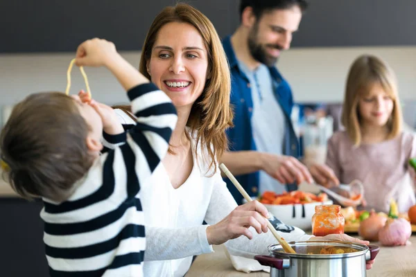 Menino bonito se divertindo com sua jovem mãe enquanto come espaguete na cozinha em casa . — Fotografia de Stock