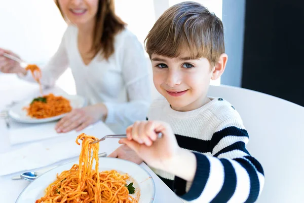 自宅のキッキングでカメラを見ながらスパゲッティを食べる陽気な小さな男の子. — ストック写真