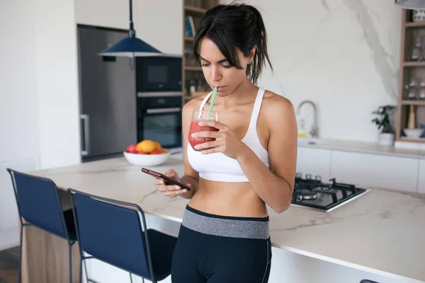 Mujer joven deportiva usando su teléfono móvil mientras bebe batido de fresa en la cocina en casa . —  Fotos de Stock