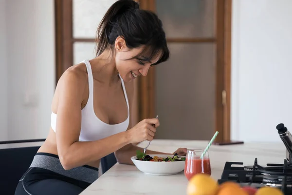 Mujer joven deportiva comiendo ensalada y bebiendo jugo de frutas en la cocina en casa . — Foto de Stock