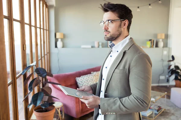 Joven hombre de negocios guapo mirando hacia los lados mientras usa su tableta digital en el pasillo de la oficina . — Foto de Stock