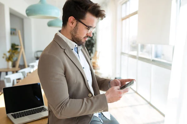 Knappe jonge zakelijke man met behulp van zijn smartphone terwijl zittend op tafel in het kantoor. — Stockfoto