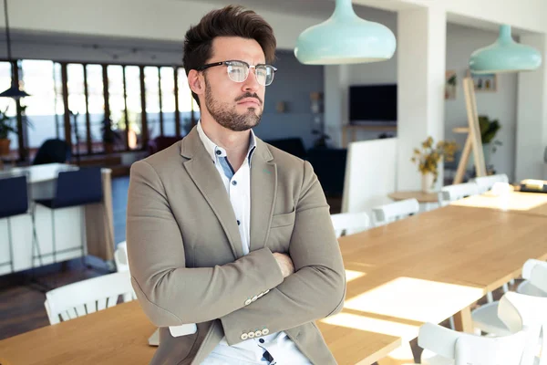 Schöner junger Geschäftsmann, der seitwärts schaut, während er im Büro auf dem Tisch sitzt. — Stockfoto