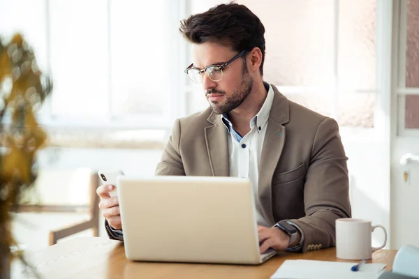 Joven hombre de negocios guapo usando su teléfono inteligente mientras trabaja con el ordenador portátil en la oficina . — Foto de Stock