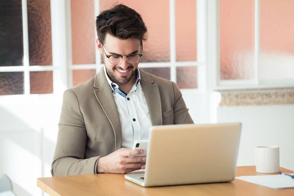 Bello giovane uomo d'affari che utilizza il suo smartphone mentre lavora con il computer portatile in ufficio . — Foto Stock