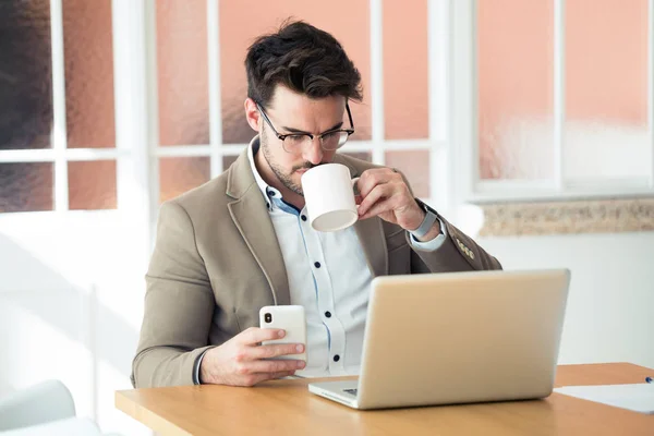 Bonito jovem homem de negócios usando seu smartphone enquanto bebe café na frente do laptop no escritório . — Fotografia de Stock