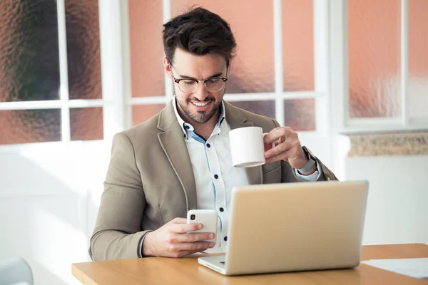 オフィスでラップトップの前でコーヒーを飲みながらスマートフォンを使うハンサムな若いビジネスマン. — ストック写真