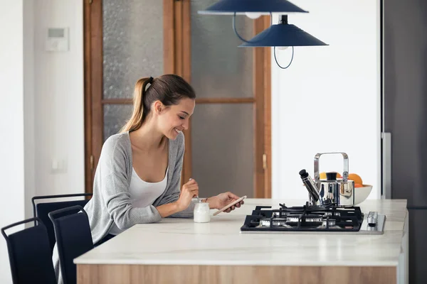 Mulher bonita enviando mensagens com telefone celular enquanto come iogurte em casa . — Fotografia de Stock