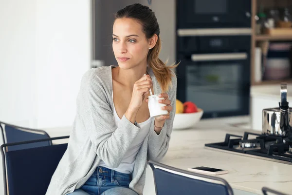 Mujer bastante joven mirando hacia los lados mientras come yogur en la cocina en casa . — Foto de Stock