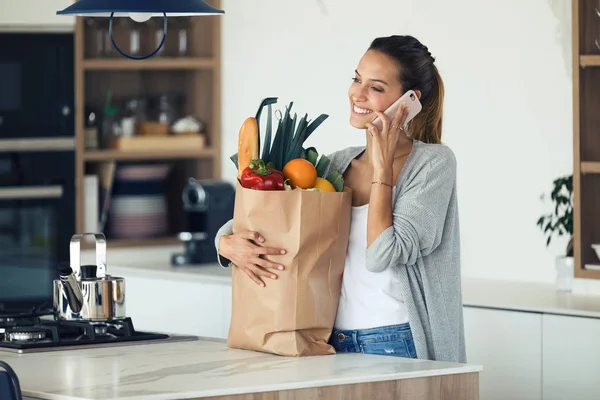 Mujer bastante joven hablando en su teléfono móvil mientras sostiene la bolsa de compras con verduras frescas en la cocina en casa . — Foto de Stock