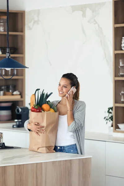 Mujer bastante joven hablando en su teléfono móvil mientras sostiene la bolsa de compras con verduras frescas en la cocina en casa . — Foto de Stock