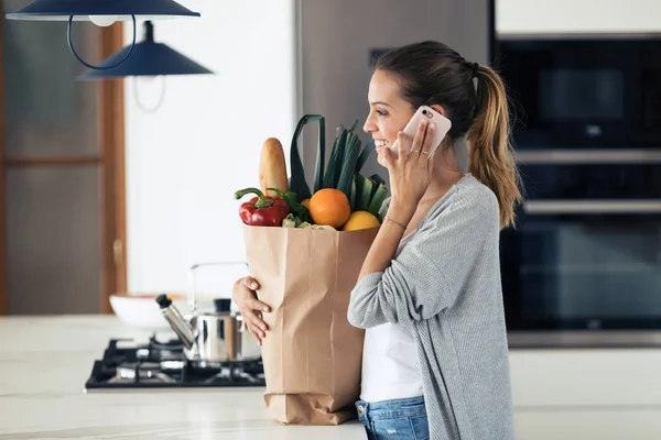 Mujer bastante joven hablando en su teléfono móvil mientras sostiene la bolsa de compras con verduras frescas en la cocina en casa . — Foto de Stock