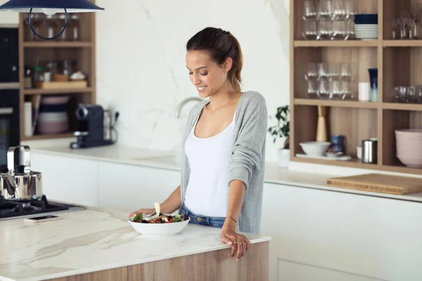 Mujer bastante joven mirando su ensalada mientras está de pie en la cocina en casa . — Foto de Stock