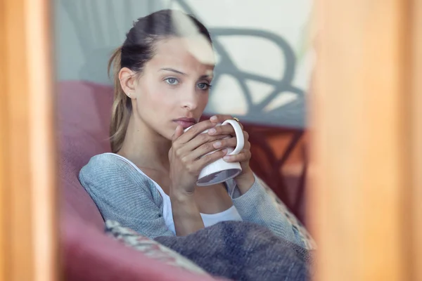 Evde kanepede kahve içerken pencereden bakan ciddi genç kadın. — Stok fotoğraf