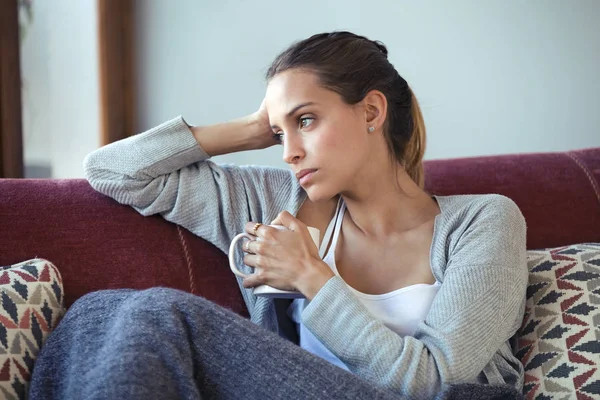 Depressive junge Frau denkt an ihre Probleme, während sie zu Hause auf dem Sofa Kaffee trinkt. — Stockfoto