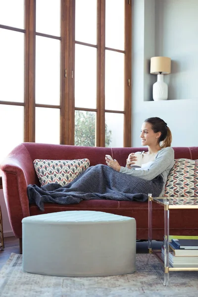 Hübsche junge Frau schaut aus dem Fenster, während sie ihr Handy zu Hause auf dem Sofa benutzt. — Stockfoto