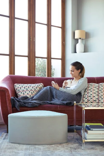 Hübsche junge Frau benutzt ihr Handy beim Kaffeetrinken zu Hause auf dem Sofa. — Stockfoto